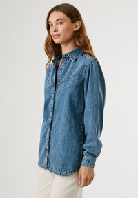 Denim košulja svijetloplave boje s prednjim kopčanjem na dugmad, šiljastim ovratnikom i dugim rukavima. Ima džepove na prsima i ležeran kroj.