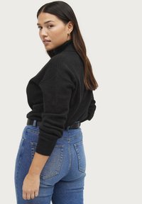 Maglione nero a collo alto, con polsini a costine e una vestibilità ampia, abbinato a jeans blu a vita alta con tasche posteriori.
