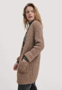 Cardigan en tricot de couleur marron clair avec une coupe ample, une ouverture sur le devant et deux poches latérales. Doté de poignets côtelés et d'un tissu doux et texturé.