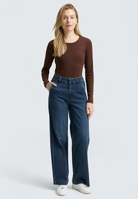Brown long-sleeve top paired with high-waisted, wide-leg blue jeans. Model wears white sneakers. Smooth textures and clean lines.
