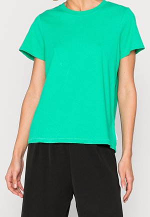 Persoon draagt een groene T-shirt met korte mouwen en losse zwarte broek, staand tegen een effen lichte achtergrond.