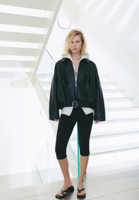 Svart läder oversized jacka med bälte, grå hoodie under, svarta leggings och remsandaler, placerad mot en ljus trappa.