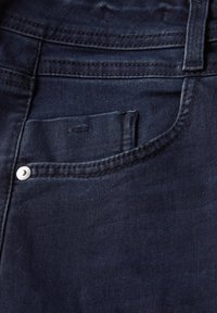 Jeans de mezclilla azul oscuro con una textura suave, que presentan un pequeño bolsillo frontal, un botón plateado y detalles de costura en contraste.
