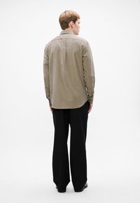 Mies, joka seisoo selin, päällään vaaleanruskea pitkähiha Shirt ja löysät mustat housut, yksinkertaista valkoista taustaa vasten.