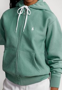 Felpa verde con zip realizzata in tessuto morbido, con un laccio bianco, dotata di tasca frontale e un piccolo logo ricamato sul petto.