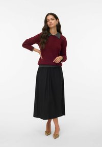 Burgunderfarbener Strickpullover mit grauem Kragen, kombiniert mit einem schwarzen, plissierten Maxirock. Das Accessoire sind gemusterte High Heels.