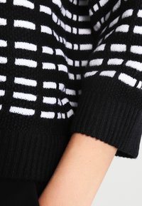 Czarnobiały dziergany sweter z wzorem w kratę, ściągaczami na rękawach i miękką teksturą. Widoczne detale dzianiny na ramionach.