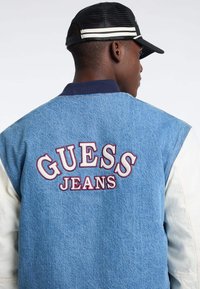 Niebieska dżinsowa kurtka z białymi skórzanymi rękawami, z haftowanym logo "GUESS JEANS" w kolorze bordowym. Żebrowany kołnierz, luźny krój.