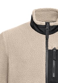 Μπεζ υφασμάτινο μπουφάν από φλις με μαύρο νάιλον γιακά και δύο κάθετα μαύρα φερμουάρ στην περιοχή του στήθους.