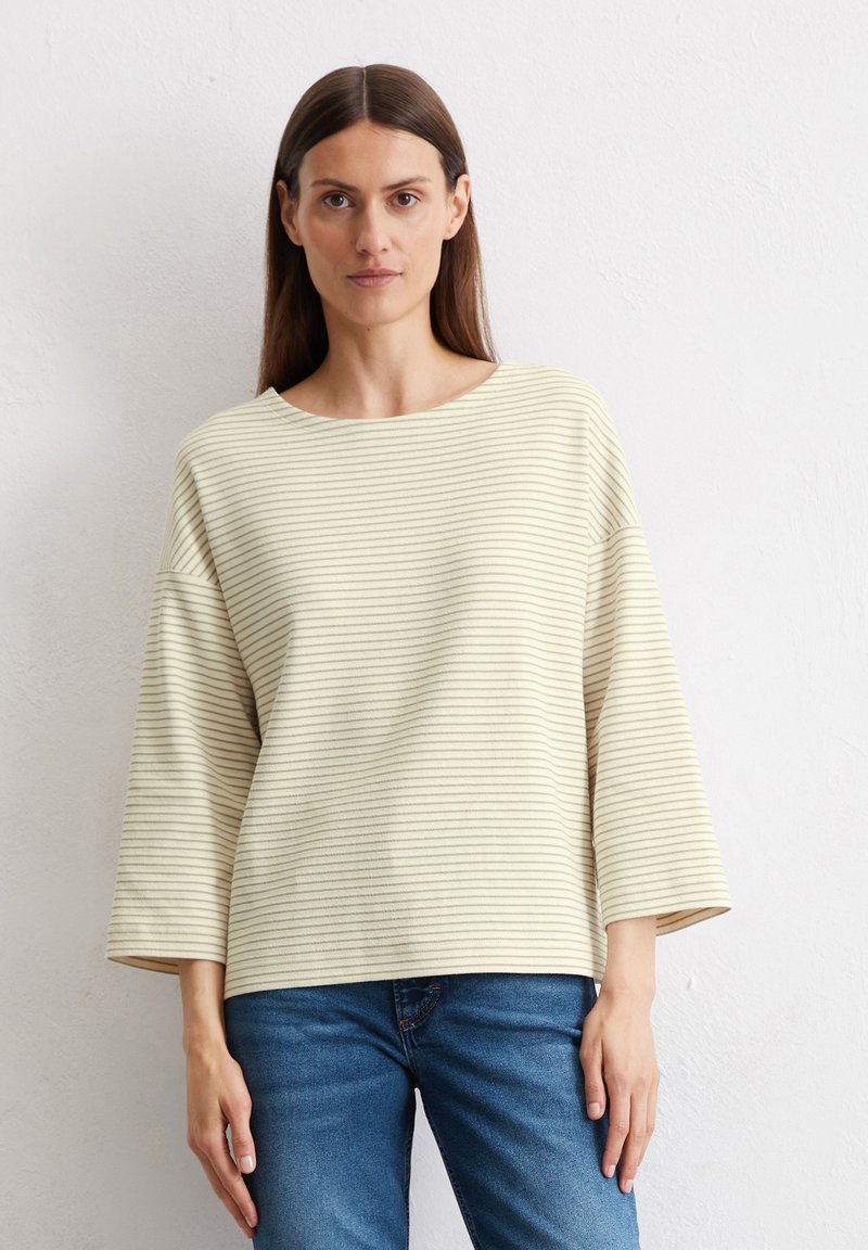 Un haut beige clair à rayures avec des manches larges et un col rond, fabriqué dans un tissu texturé. Associez-le à un jean bleu ajusté.