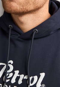 Námořnická modrá mikina s kapucí, s přední kapsou, šňůrkami na stažení a s texturovaným potiskem loga. Obsahuje kovové průchodky a měkký povrch látky.