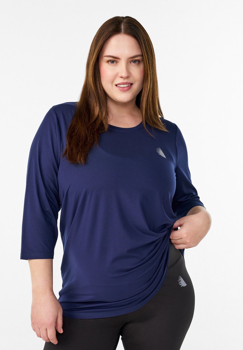 Navyblaues Langarmshirt aus weichem Stoff mit rundem Ausschnitt und dezentem Logodetail. Kombiniert mit dunklen Leggings und präsentiert einen entspannten Sitz.