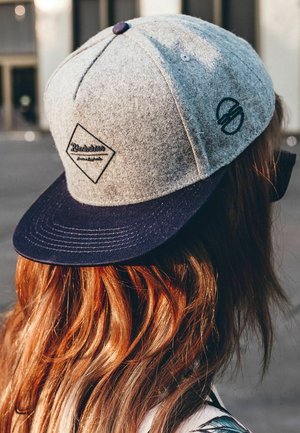 Grauem Snapback-Cap mit einem navyfarbenen Schirm, das ein rautenförmiges Logo in Schwarz und einen runden Akzent an der Seite zeigt, getragen mit welligem Haar.