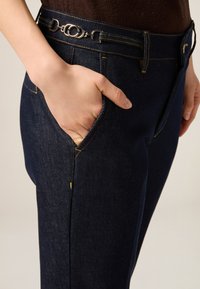 Pantalones de mezclilla azul marino con herrajes plateados, trabillas para cinturón y costuras prominentes. Cuentan con un bolsillo lateral y un diseño de corte slim.