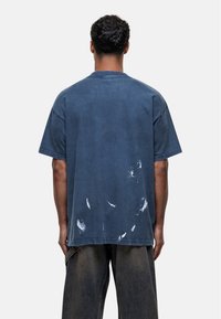 T-shirt azzurro in cotone oversize con maniche corte, caratterizzato da una finitura consumata e schizzi bianchi simili alla vernice sul retro.