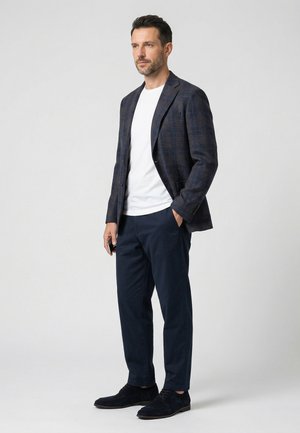 Muž stojící, oblečený v tmavém kostkovaném saku, bílém tričku, tmavě modrých kalhotách a černých botách, drží v jedné ruce telefon.