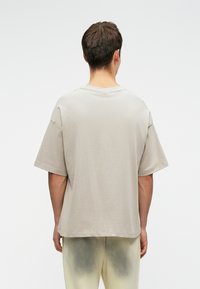 Tricou din bumbac gri deschis, oversized, cu mâneci scurte, guler rotund, cusături vizibile pe umeri, potrivire lejeră. Vedere din spate.