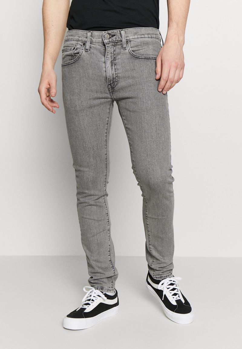 Homme portant un jean gris slim et des baskets noir et blanc, debout contre un mur blanc uni.