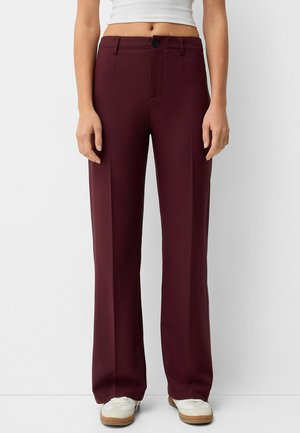 Woman wearing high-waist, wide-leg burgundy trousers with front crease, black button, paired with white sneakers and cropped white top.