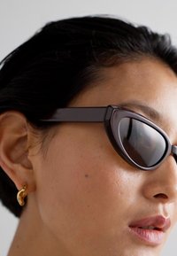 Cat-eye solbriller med mørkt tonede linser, mat sort stel, vinklet design og en subtil guld hoops earring synlig i øret.