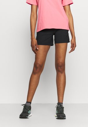 Henkilö, jolla on vaaleanpunainen lyhythihainen paita, mustat shortsit ja mustavihreät urheilukengät, seisoo kasvot eteenpäin yksiväristä taustaa vasten.