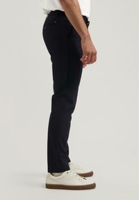 Pantalon droit noir en tissu durable, avec poches latérales et taille à boutons. Associé à des baskets blanches.