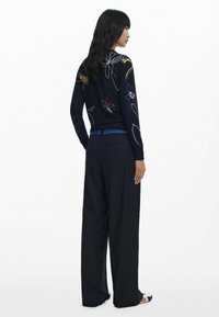 Marineblauer Pullover mit buntem Blumenstickerei, kombiniert mit weit geschnittenen schwarzen Hosen. Die Hosen haben einen blau akzentuierten Bund und Taschen.