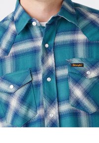 Blauwe en teal geruite flanellen overhemd met twee knoopzakken op de borst, witte hardware en een Wrangler-logo op de linker zak.