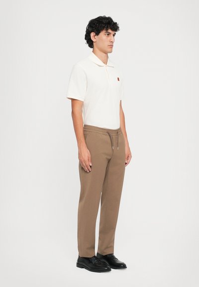 Cremefarvet polo skjorte med korte ærmer og et lille logo, parret med brune bukser med elastisk talje og sorte sko, stående foran en ensfarvet baggrund.