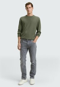 Sweter oliwkowozielony z prążkowanymi mankietami, w połączeniu z szarymi dżinsami i białymi tenisówkami. Prosty fason, dopasowana sylwetka, minimalne oznaczenia na swetrze.