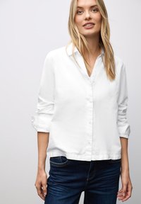 Mujer rubia con una camisa blanca de botones y mangas arremangadas, y jeans azul oscuro, de pie contra un fondo gris claro.