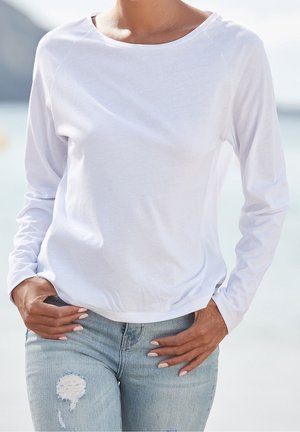 Kobieta ubrana w gładką białą bluzkę z długim rękawem i jasnoniebieskie podarte dżinsy, stojąca z rękami w kieszeniach na tle rozmytego, zewnętrznego tła.