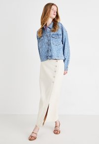 Veste en denim bleu clair avec deux poches sur la poitrine, associée à une jupe beige à boutons, arrivant au genou, avec une fente sur le côté. Sandales à lanières marron.