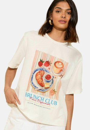 Frau trägt ein weißes T-Shirt mit einer Brunch-Themen-Illustration, die ein Croissant, Kaffee, Erdbeeren und den Text "Brunch Club Petit-Dejeuner" zeigt.