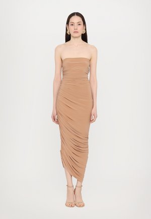 Vestido beige ajustado sin tirantes con textura fruncida, de longitud hasta la rodilla y abertura lateral, acompañado de tacones con tiras. Acentuado con grandes pendientes.