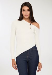 Kobieta w białym swetrze typu one-shoulder z prążkowanej tkaniny i ciemnoniebieskich dżinsach, stojąca na tle jasnego, gładkiego tła.
