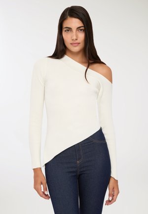 Donna che indossa un maglione bianco a spalla scoperta con coste e jeans blu scuro, in piedi contro uno sfondo chiaro e semplice.