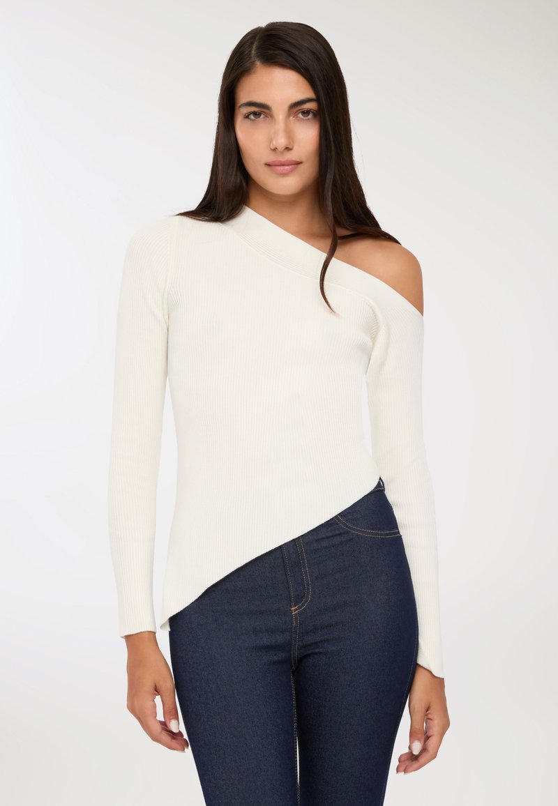 Kobieta w białym swetrze typu one-shoulder z prążkowanej tkaniny i ciemnoniebieskich dżinsach, stojąca na tle jasnego, gładkiego tła.