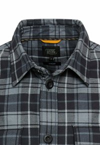 Chemise overshirt à carreaux gris et noir avec un col à boutons, deux boutons à l'avant et une poche poitrine. Étiquette visible à l'intérieur du col.