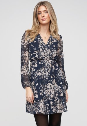 Młoda kobieta o długich blond włosach ubrana w granatową sukienkę w kwiaty z długimi, przezroczystymi rękawami oraz czarnymi rajstopami, stoi na tle gładkiej ściany.