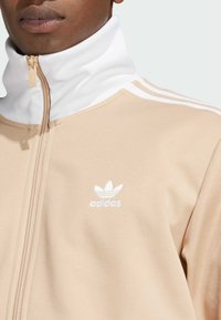 Bež jakna na zip s visokim ovratnikom, bijelim detaljima i tri bijele pruge na ramenima. Na njoj se nalazi bijeli Adidas logo.