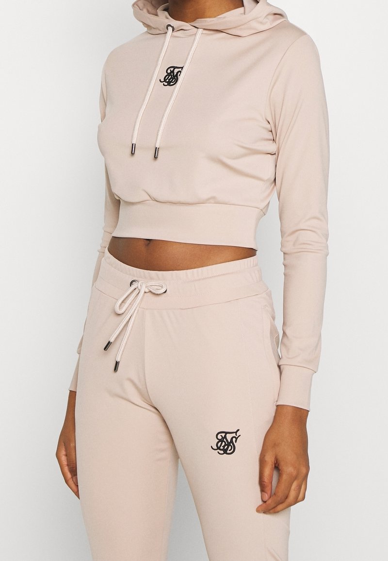 Beige verkürzter Kapuzenpullover mit Kordelzug und Logo auf der Brust, kombiniert mit passenden, eng anliegenden Jogginghosen mit elastischem Bund und Logoakzent.