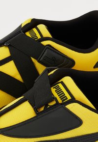 Baskets Puma jaunes et noires avec tige en mesh, brides croisées élastiques noires et logo Puma sur la languette et les détails des brides.