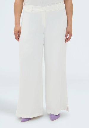 Femme portant un pantalon blanc taille haute à jambes larges associé à des chaussures pointues lavande, debout devant un arrière-plan clair uni.