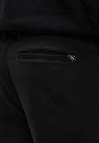 Κοντινό της πίσω τσέπης μαύρων παντελονιών με μια μικρή ετικέτα λογότυπου Puma στην επάνω δεξιά γωνία της τσέπης.