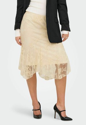 Femme portant une jupe midi en dentelle beige, un blazer noir, un pull en maille blanche côtelée et des chaussures Mary Jane noires à talons hauts, debout sur un fond clair.