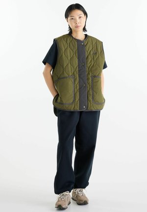 Giovane in piedi che indossa un gilet imbottito verde oliva sopra una camicia nera, pantaloni neri e sneakers beige su sfondo bianco.