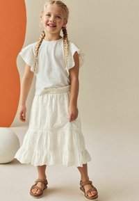 Haut blanc avec manches en dentelle, jupe blanche brodée, ourlet volanté et sandales marron. Design simple sur un fond neutre.
