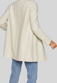 Abrigo de crema texturizado con un corte relajado, mangas largas y longitud por debajo de la cadera. Presenta bolsillos laterales y un diseño de cuello minimalista.