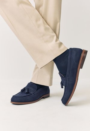 Mocassins à pampilles bleu marine portés avec des chaussettes bleu marine et un pantalon beige, montrant un pied levé et croisé sur l'autre.